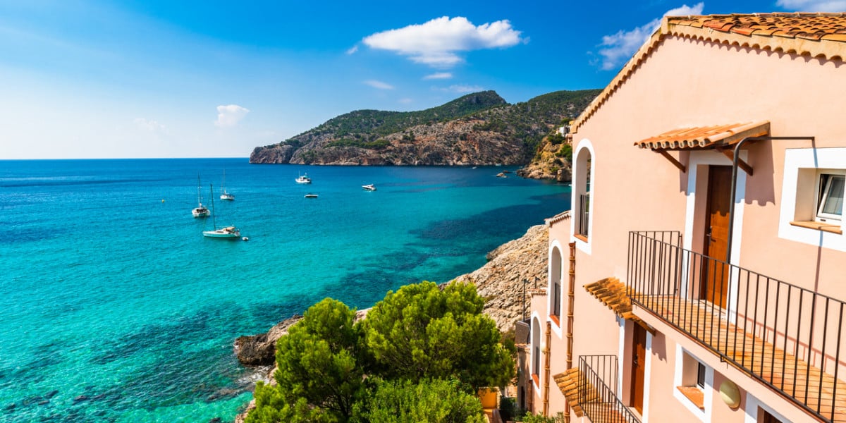 Vista de una costa mediterránea con aguas turquesas, barcos anclados y casas de colores pastel que dominan el mar bajo un cielo azul despejado