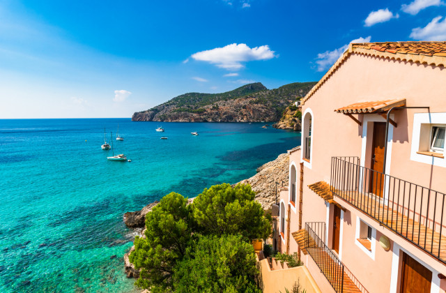 Vista de una costa mediterránea con aguas turquesas, barcos anclados y casas de colores pastel que dominan el mar bajo un cielo azul despejado