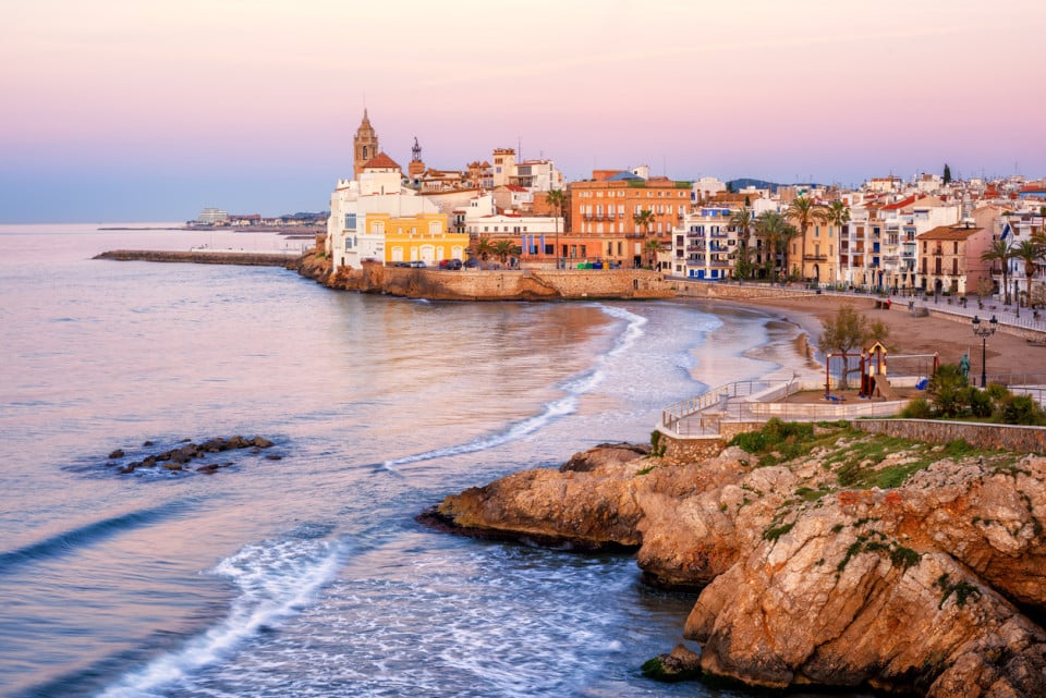 Atardecer en la costa de Sitges, con mar tranquilo que refleja tonos dorados y naranjas, y casas mediterráneas tradicionales alineadas a la costa